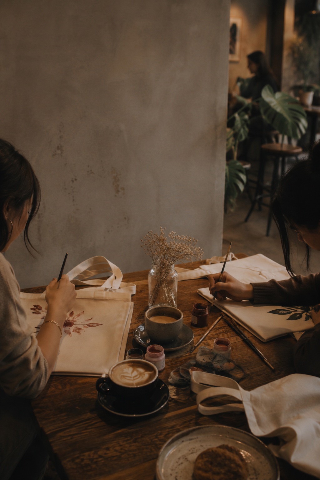 Two women painting floral designs on canvas tote bags at a warm café table